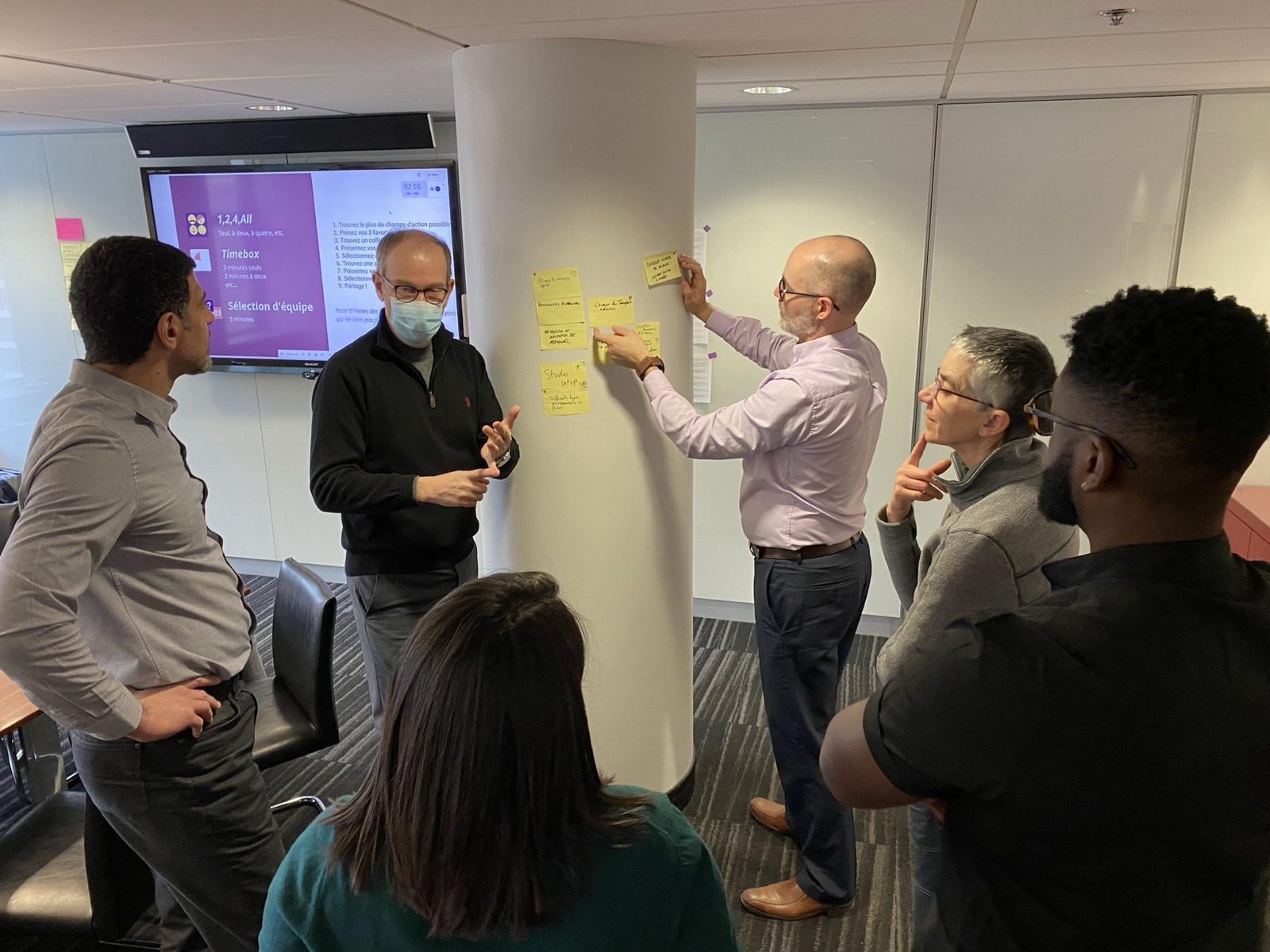 Des participants rassemblés autour d’une colonne couverte de post-it pendant qu’un d’eux en ajoute un