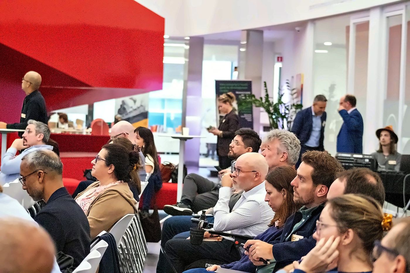 Attendees listening to a talk in the main hall at Espace Ax.C, under the red staircase